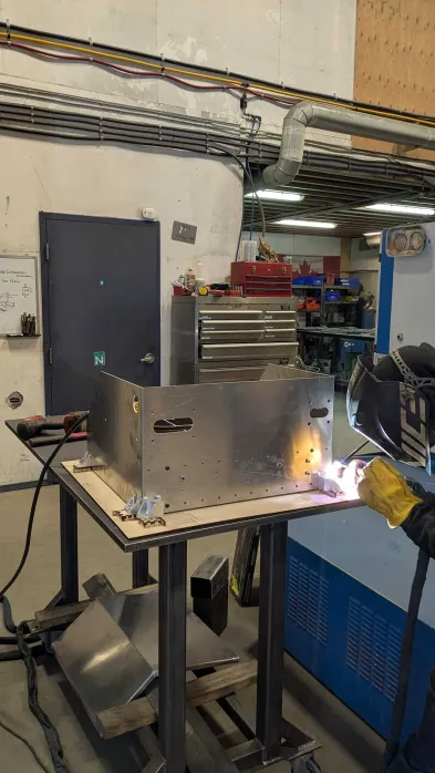Teammate TIG welding on the aluminum accumulator enclosure, clamped to the jig baseplate